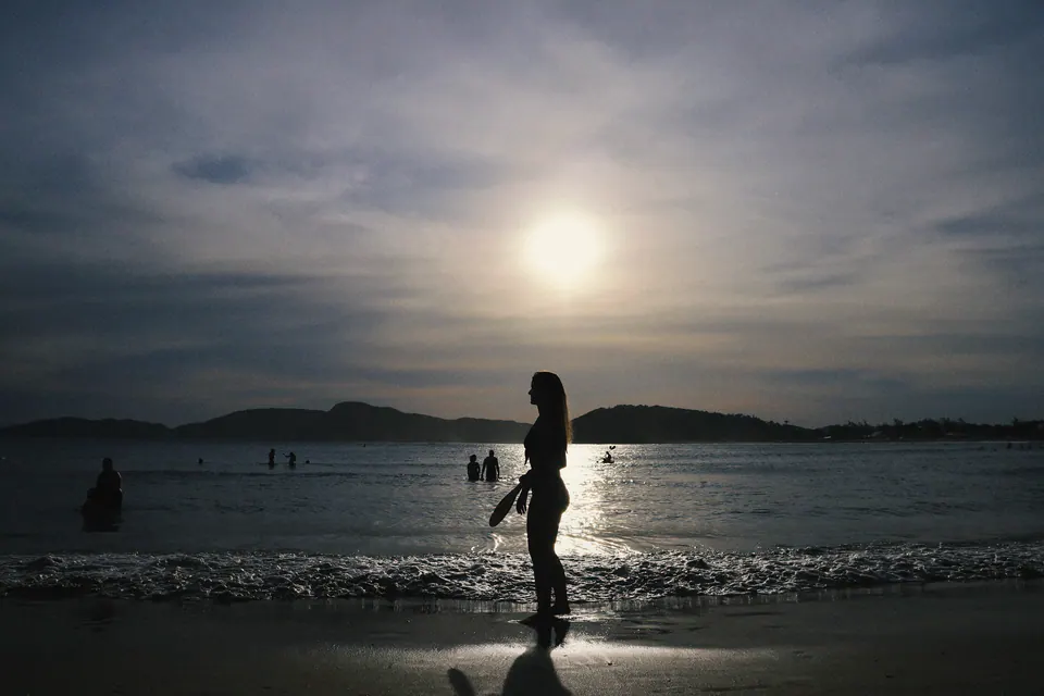 silhueta de mulher na beira do mar contra o sol, com o reflexo na areia molhada